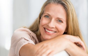 A smiling woman with long blonde hair rests her arms on a surface, looking at the camera., 