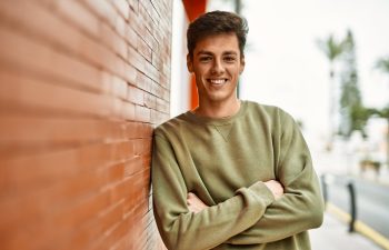 Person in a green sweater leaning against a brick wall, smiling with arms crossed, outdoor setting., 